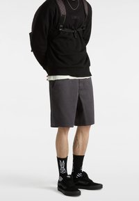 Felpa nera, pantaloncini grigi, sneaker nere e calzini neri decorati con coniglietti bianchi e mani scheletriche. Zaino con motivo a scacchiera.