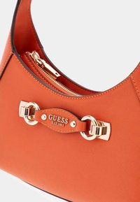 Pomarańczowa syntetyczna torba o zaokrąglonym kształcie, z złotymi akcentami, zapięciem na zamek oraz skórzaną metką z logo. Teksturowana powierzchnia.