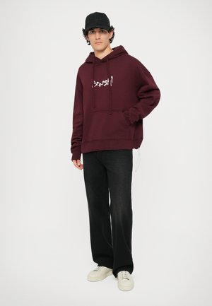 Haut de sweatshirt bordeaux avec texte blanc, doté d'une poche kangourou et d'une capuche avec cordon de serrage, associé à un pantalon large noir et des baskets blanches.