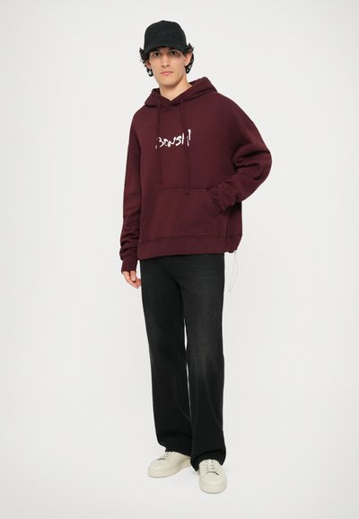 Burgundin pullover huppari valkoisella tekstillä, jossa on kengurupocket ja narukiinnitys, yhdistetty mustiin leveälahkeisiin housuihin ja valkoisiin tenniskenkiin.