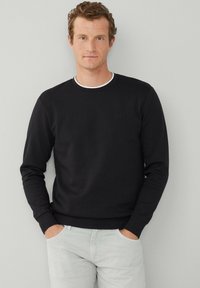 Sweat-shirt noir avec un col rond, doté d'un col intérieur blanc. Texture douce, manches longues et poignets côtelés, associé à un pantalon gris clair.