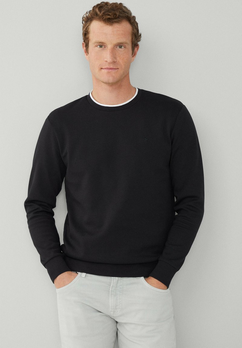 Sweat-shirt noir avec un col rond, doté d'un col intérieur blanc. Texture douce, manches longues et poignets côtelés, associé à un pantalon gris clair.