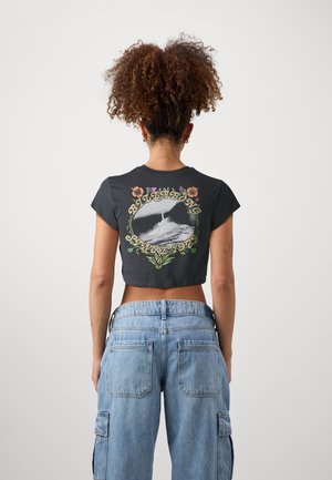 Frau mit lockigem Haar, die ein schwarzes Croptop mit Wellendesign und Text trägt, kombiniert mit hellblauen, hoch taillierten Cargohosen.