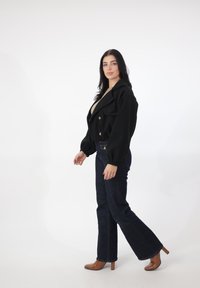 Sort cropped dobbeltradet jakke med guldknapper, kombineret med mørke, flarede jeans og brune sko med hæl, sat mod en hvid baggrund.