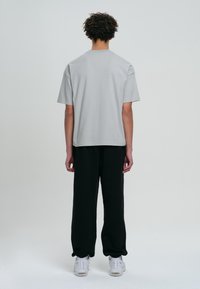 Jasnoszara koszulka z krótkim rękawem, luźny krój, zestawiona z czarnymi spodniami. Model stoi tyłem, na białej podłodze, w białych trampkach.