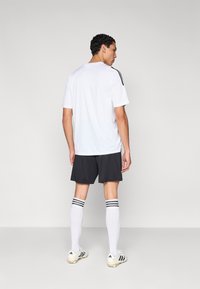 Bijela sportska majica s crnim prugama na ramenima, uparena s tamnim kratkim hlačama, bijele čarape do koljena s crnim prugama i kopačke.