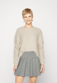 Flauschiger, cremefarbener Pullover mit langen Ärmeln und rundem Ausschnitt, kombiniert mit einem grauen, plissierten Rock mit kariertem Muster.