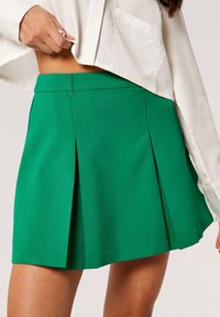 Grüne Faltenrock aus einem glatten, strukturierten Stoff, hat eine enganliegende Taille mit einem Reißverschluss. Der Saum endet auf halber Oberschenkelhöhe.