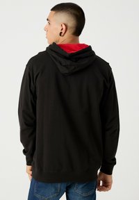 Sudadera negra con un forro rojo en el interior de la capucha, tela texturizada, mangas largas y un ajuste relajado, combinada con jeans azules.