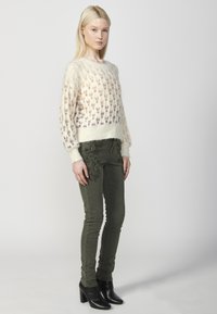 Pulover tricotat crem cu modele florale în roz și mov; blugi ajustați de culoare verde măsliniu cu detalii florale brodate; cizme negre cu toc.