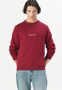 Burgundy sweatshirt made of cotton, with long sleeves and crew neck. Features small white embroidered logo on the chest. Worn with light blue jeans.