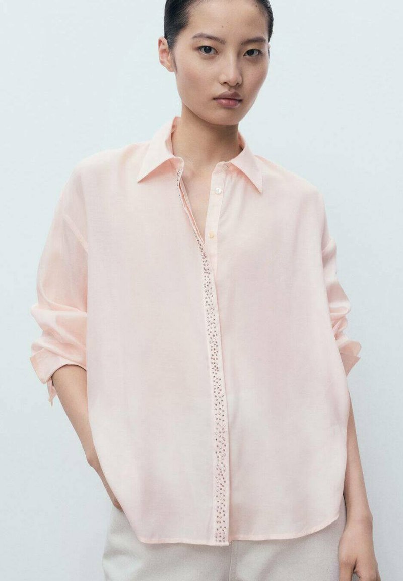 Camicia button-up color pesca chiaro realizzata in tessuto trasparente. Presenta maniche arrotolate, colletto classico e bottoni decorativi lungo la patta.