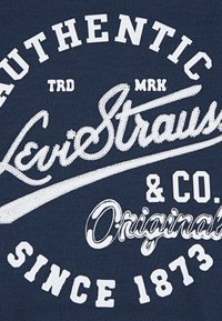 Mörkblå tyg t-shirt med vit broderad text: "AUTENTISKA Levi Strauss & Co. Original sedan 1873" som har dekorativ sömnad.