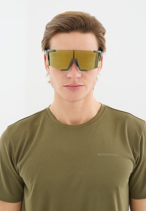 Jonge man met grote, hoekige, geel getinte sportzonnebril en een olijfgroen T-shirt met de tekst "ENDURANCE" op de borst.