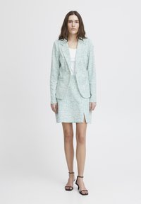 Světle modrý vzorovaný blazer s odpovídající sukní, která má rozparek. Tkanina má hladkou texturu, doplněná o černé sandály na řemínky.