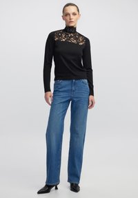 Zwarte langemouwen top met een kant high neck, gecombineerd met blauwe wijde jeans en zwarte puntige hakken. Simpel en elegant ontwerp.