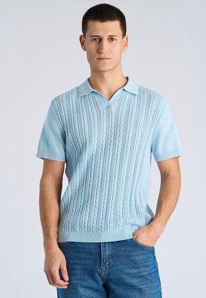 Jonge man kijkt naar voren met kort bruin haar, draagt een lichtblauw gebreid polo met korte mouwen en een blauwe spijkerbroek, met één hand in de zak.