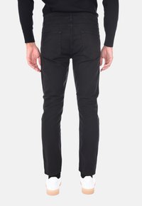 Persona che indossa jeans neri e sneakers bianche, mostrata di spalle su uno sfondo chiaro e semplice.