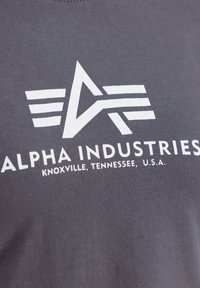 Grå bomulls t-skjorte med et stort hvitt logo med et abstrakt design og teksten "ALPHA INDUSTRIES, KNOXVILLE, TENNESSEE, U.S.A."