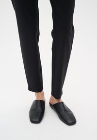 Černé kožené slip-on muly s designem čtveraté špičky, spárované s elegantními černými kalhotami. Muly mají hladký povrch a minimální zdobení.