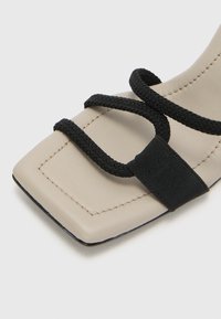 Beige Flachsandale mit quadratischer Zehenpartie, ausgestattet mit schwarzen geflochtenen Riemen und einem elastischen Akzent. Glatte Textur mit kontrastierenden Nähten.