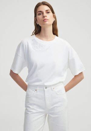 T-shirt en coton blanc avec un col rond, des manches courtes et des détails en dentelle florale sur l'épaule gauche et la manche. Associé à un pantalon blanc.