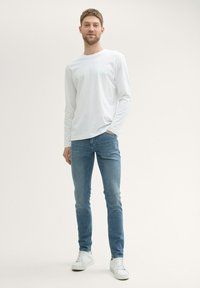 Camiseta blanca de manga larga, jeans ajustados azul oscuro y zapatillas blancas. Diseño minimalista con textura suave y sin patrones o logotipos visibles.