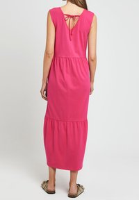 Rosa Baumwoll-Maxikleid mit einem taillierten Oberteil, schichtartigem Rock und einem Schlüsselloch-Rücken mit einer Schleife gebunden. Flache Sandalen vervollständigen den Look.