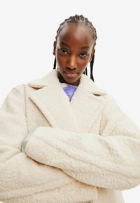 Kreemikas, pehme ja karvane mantel kahe rea nööpidega, suurte kraede ja volditud varrukatega. Tekstuuriga kangas, millel on pehme viimistlus.