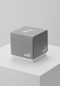 Boîte Puma grise et blanche avec logo sur le dessus et les côtés, placée sur un piédestal blanc devant un fond gris clair.