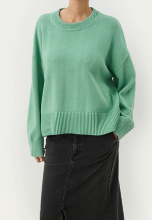 Personne portant un pull en maille vert menthe ample et une jupe en denim noire taille haute avec une fente devant, debout avec une main dans la poche.