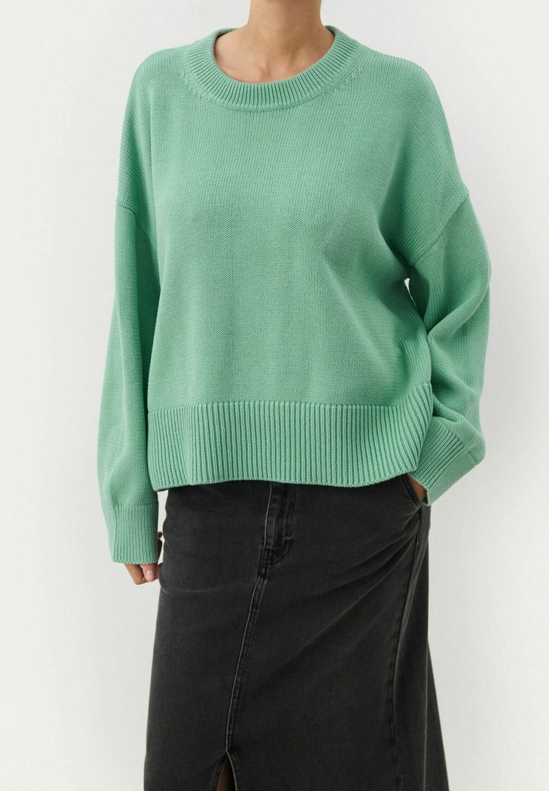 Personne portant un pull en maille vert menthe ample et une jupe en denim noire taille haute avec une fente devant, debout avec une main dans la poche.