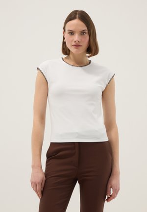 Femme aux cheveux courts bruns portant un haut blanc à manches courtes avec bordure noire et un pantalon marron taille haute, debout devant un fond uni.