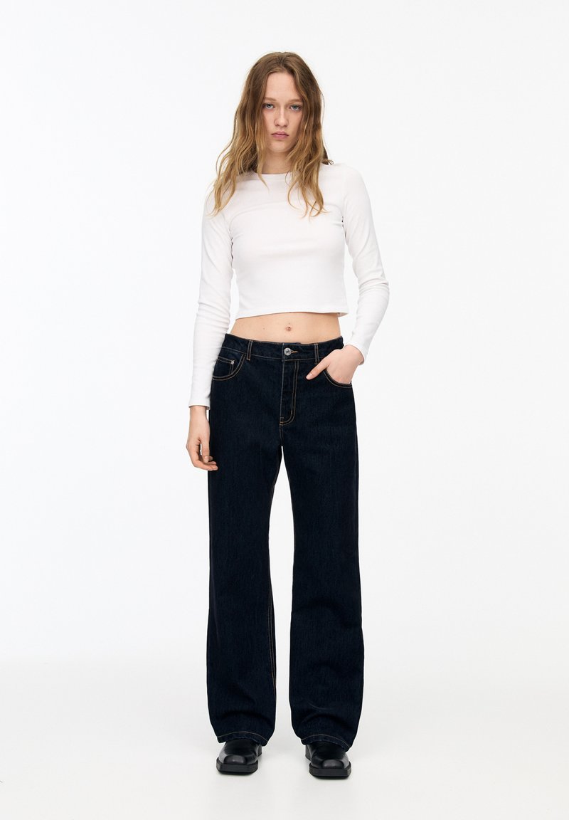 Weißes, kurz geschnittenes Longsleeve-Oberteil und hochgeschnittene, dunkle Jeans mit weitem Bein und kontrastierenden Nähten sowie Vorderpockets, getragen mit schwarzen Schuhen.