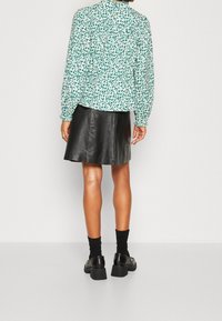 Blumenbluse mit grünem und weißem Muster, kombiniert mit einem schwarzen Lederrock. Modell trägt schwarze Socken und klobige schwarze Schuhe.