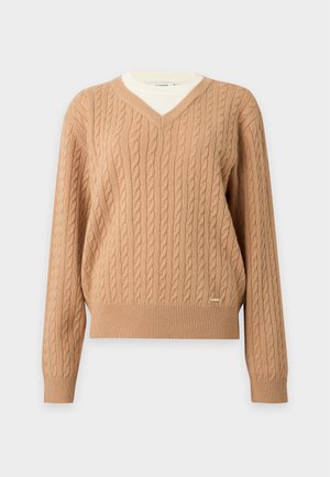 Femme portant un pull en maille torsadée beige, une chemise blanche, un pantalon marron ceinturé et des mocassins noirs, debout devant un fond clair uni.