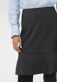 Jupe grise foncée jusqu'au genou en tissu texturé, présentant un design superposé avec une finition lisse. La main du modèle repose sur le tissu.