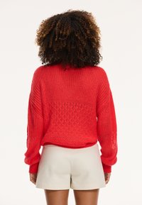 Maglione rosso lavorato a maglia con un motivo testurizzato e una vestibilità ampia, caratterizzato da maniche larghe e una lunghezza cropped, abbinato a pantaloni corti color crema.