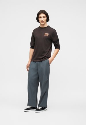 Noor mees keskmise pikkusega tumedate juustega, kandes musta pikavarukalist graafilise mustriga särki, lahtisi hallid pükse ja musti tossusid valgete talladega.