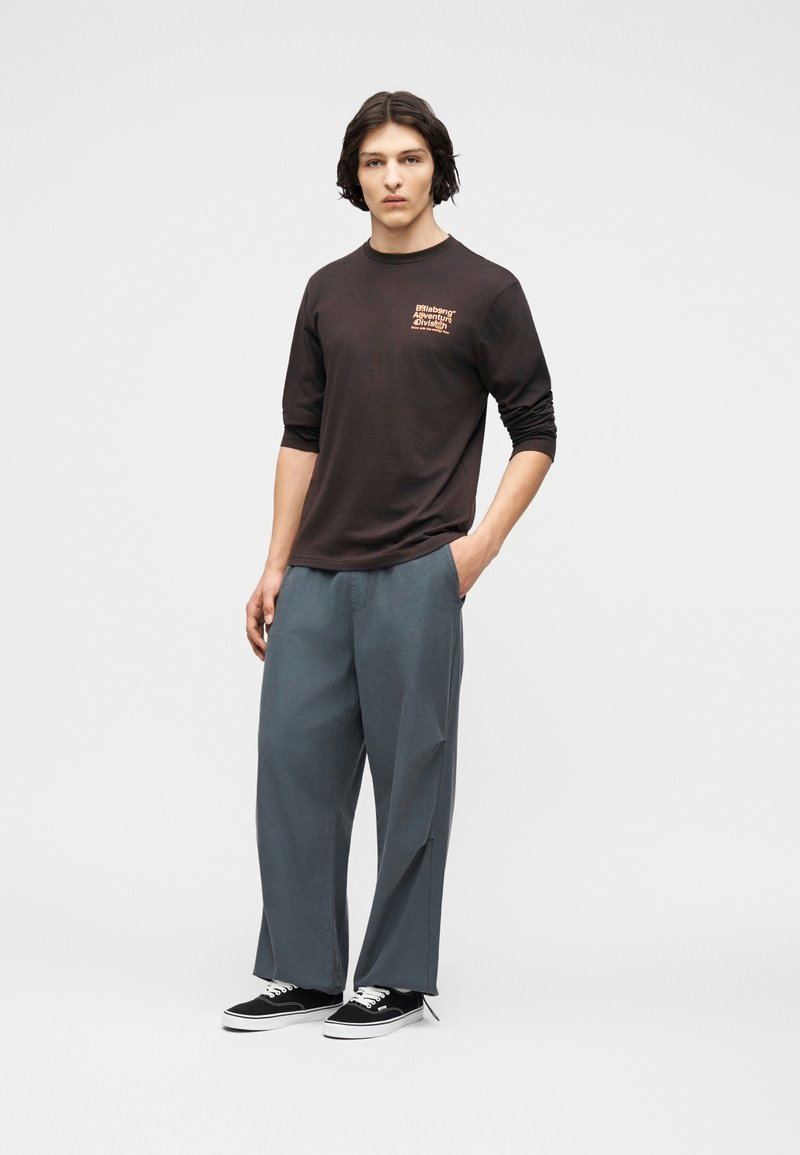 Νεαρός άνδρας με μεσαίου μήκους σκούρα μαλλιά, φοράει μαύρο μακρυμάνικο πουκάμισο με σχέδιο, φαρδιά γκρι παντελόνια και μαύρα αθλητικά παπούτσια με λευκές σόλες.