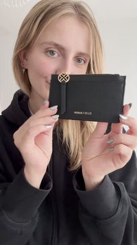 Melnās kartes turētājs ar teksturētu virsmu, zelta logo "ANNA FIELD" un dekoratīvu cilpiņu. Kompakta uzbūve ērtai uzglabāšanai.