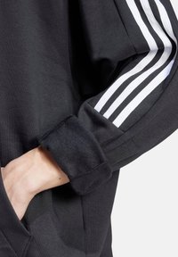 Felpa nera con una texture morbida, caratterizzata da tre strisce bianche lungo la manica e un polsino ripiegato dove è visibile una mano.