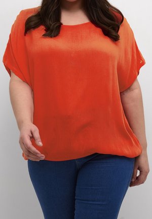 Woman wearing a loose-fitting bright orange short-sleeve blouse and dark blue jeans, standing against a plain light background.