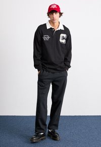 Sort langærmet polo shirt med hvid krave og bogstavet "C" som grafik, parret med sorte bukser og sorte sko med tykke såler. Rød kasket.