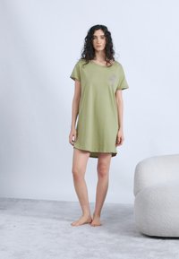 Νεαρή γυναίκα με σγουρά, σκούρα μαλλιά στέκεται ξυπόλυτη, φορώντας ένα φαρδύ φορεματάκι T-shirt σε ελιά πράσινο χρώμα με μικρό σχέδιο φοίνικα.