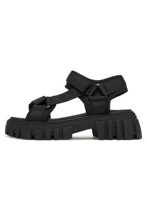 Sandalias negras con suela de goma, con tiras ajustables de tela. Suela gruesa con estrías texturizadas para mayor agarre. Diseño moderno y minimalista.