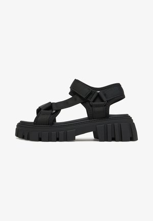 Black sandals with a rubber sole, featuring adjustable fabric straps. Chunky sole with textured ridges for grip. Modern, minimal design.