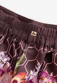 Stiept elastīga josta uz tumša auduma ar rozā ģeometrisku rakstu un krāsainu ziedu dizainu, papildināta ar mazu bēša pogu ar reljefu logo.