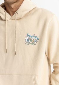 Sudadera color crema con un gráfico en el pecho que presenta flores y un personaje, acentuada con azul claro y verde. Material de algodón.