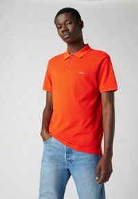 Camisa polo naranja con cuello, tres botones y logo bordado de "París". Llevada con jeans de color azul claro, mostrando un ajuste relajado.